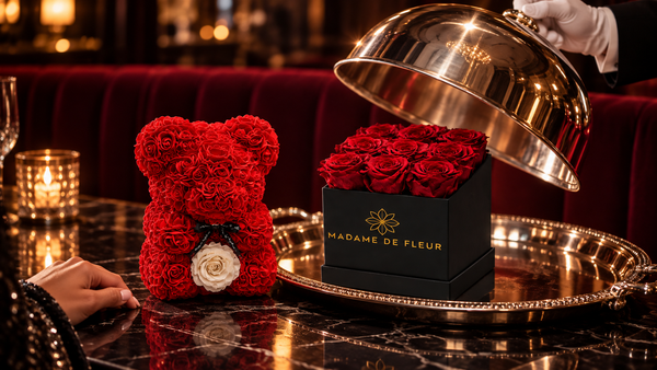 A red rose bear beside a black MADAME DE FLEUR rose box beneath a lifted silver cloche on a candlelit restaurant table.