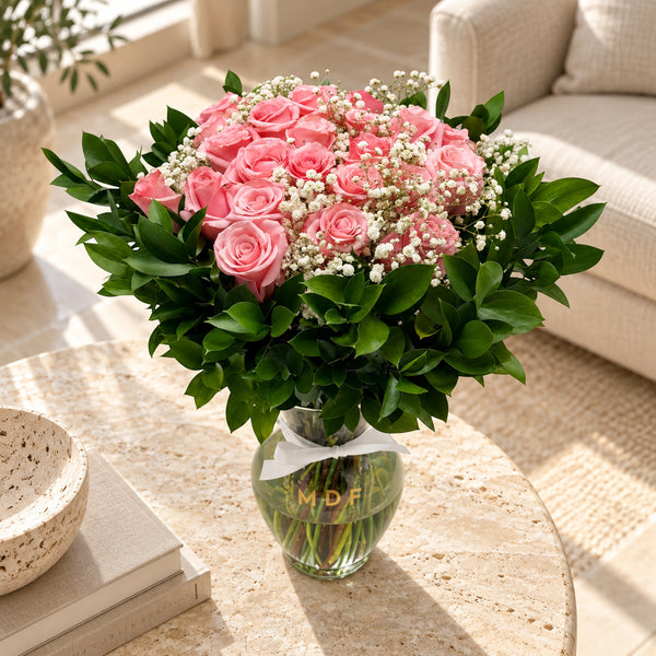 Classic 24 pink fresh roses with greenery and baby's breath in a clear glass vase, sunlit home lifestyle view