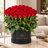 Red fresh roses with tall stems and green foliage in a black MADAME DE FLEUR box on a living-room table.